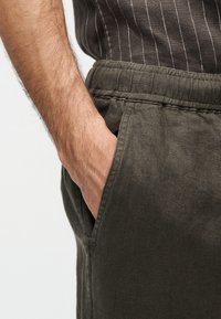 Ανδρικό χέρι που ακουμπά στην εμπρόσθια τσέπη σκούρου γκρι παντελονιού με ελαστική μέση, φοράει σκούρο πουκάμισο με λεπτές λευκές ρίγες.