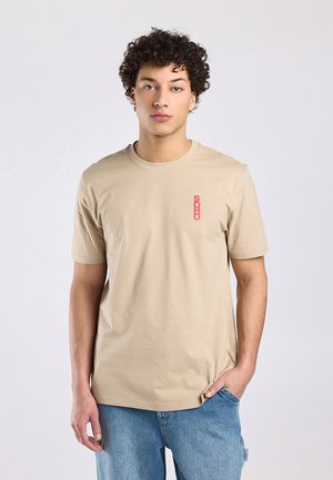 Jonge man met krullend haar, die een beige T-shirt draagt met rode "SOHO"-tekst en lichtblauwe spijkerbroek, staand met één hand in zijn zak.