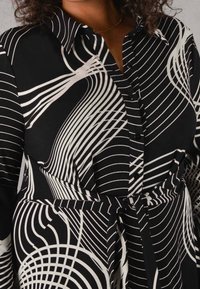 Abito camicia bianco e nero con collo a camicia, caratterizzato da motivi a strisce swirl, bottoni sul davanti e cintura annodata in vita.