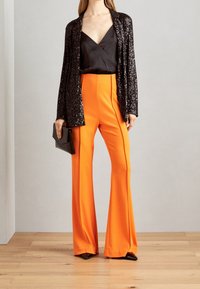 Chaqueta de punto con lentejuelas negras sobre un top de satén negro y pantalones anchos de color naranja brillante. La modelo sostiene un pequeño bolso de mano negro. El suelo es de madera.