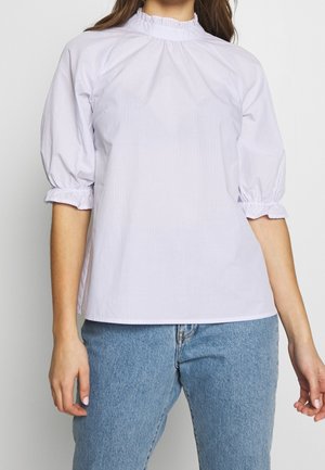 Kobieta nosząca jasnofioletową, paskowaną bluzkę z marszczonym wysokim kołnierzykiem i trzyczęściowymi bufkami, w połączeniu z niebieskimi jeansami.