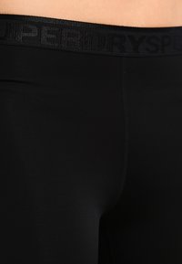 Pánevní a horní část stehna v černých sportovních legínách s černým pasem, na kterém je decentní značení "Superdry Sport".