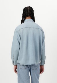 Jachetă din denim de culoare bleu deschis, cu o croială relaxată, mâneci lungi și guler clasic; are cusături vizibile și manșete cu nasturi.