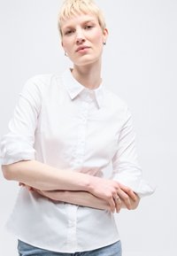 Νεαρό άτομο με κοντά ξανθά μαλλιά, φοράει ένα λευκό πουκάμισο με κουμπιά και τζιν, με σταυρωμένα τα χέρια, μπροστά σε απλό φωτεινό φόντο.