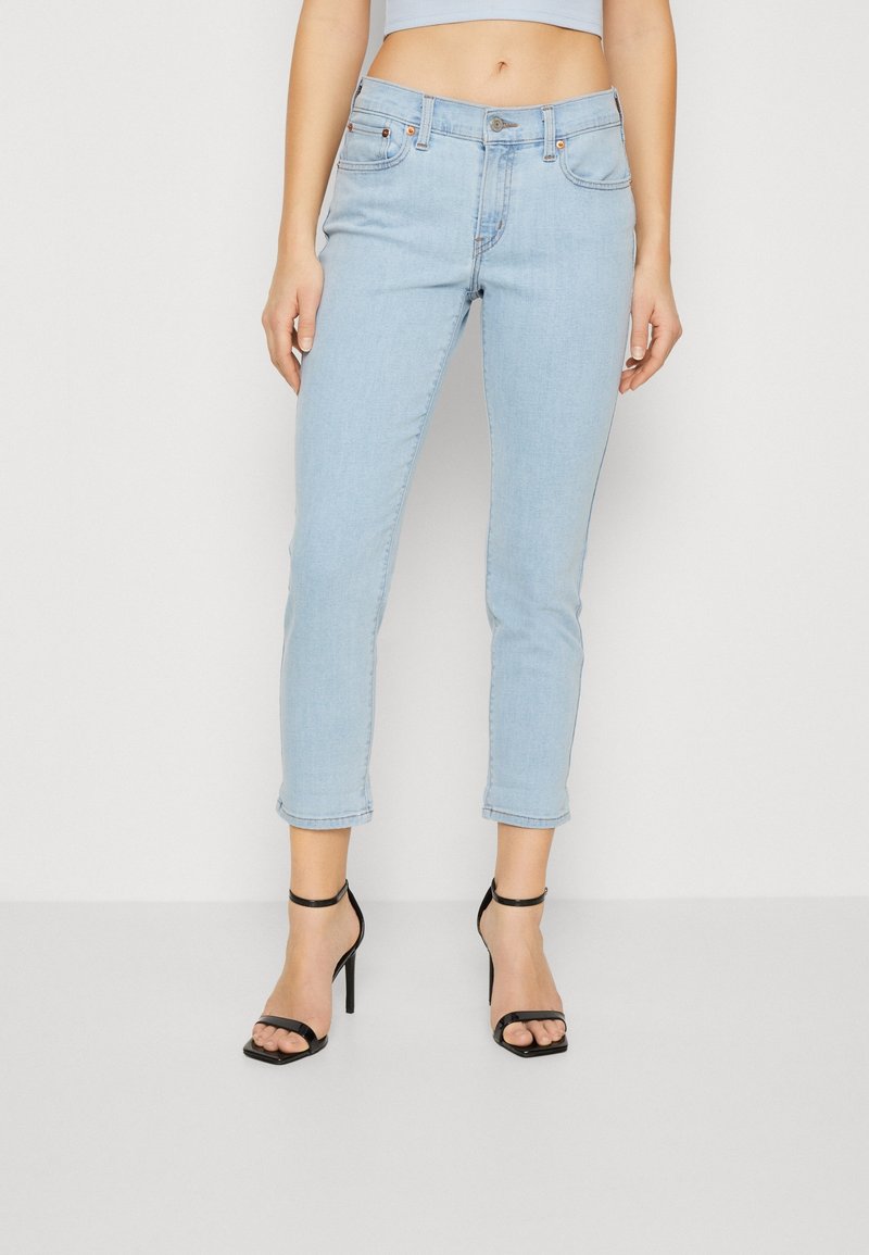 Light blue denim jeans with a slim fit, cropped length, and rolled cuffs. Features front pockets and a button closure. Paired with black strappy heels.