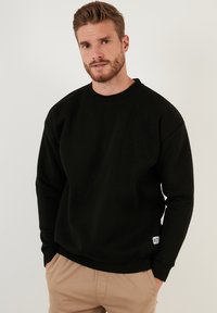 Sweatshirt negru dintr-un material moale, cu guler rotund, mâneci lungi și manșete ribbed. Include o etichetă mică la tiv.
