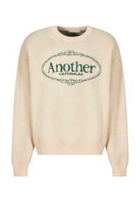 Beżowy sweter dziani z okrągłym wzorem logo z napisem "Another" w ciemnozielonym kolorze, otoczony kwiatowym motywem. Żebrowany dekolt w kształcie łódki oraz mankiety.