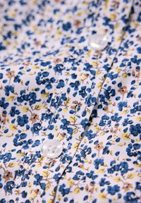 Tessuto bianco con un motivo floreale che presenta fiori blu e gialli. Un bottone trasparente è visibile vicino al centro con una texture liscia.