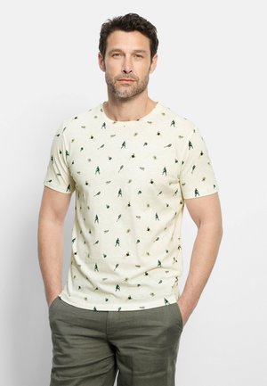 Uomo con barba che indossa una maglietta beige chiaro con motivo giocatore di tennis verde e racchetta, e pantaloni verde oliva, mani nelle tasche.