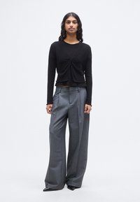 Μαύρο τοπ με μακριά μανίκια και λεπτομέρεια δέσιμου στο κέντρο, συνδυασμένο με γκρι παντελόνες φαρδιές στο πόδι. Λείο ύφασμα με διακριτικές υφές.