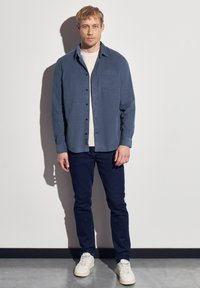 Button-up shirt in dark blue, lightweight fabric, long sleeves, chest pocket. Paired with dark jeans and white sneakers. Plain background.