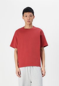 Tricou roșu din bumbac cu o croială lejeră, guler rotund și mâneci scurte, combinat cu pantaloni de trening gri deschis cu șnur.