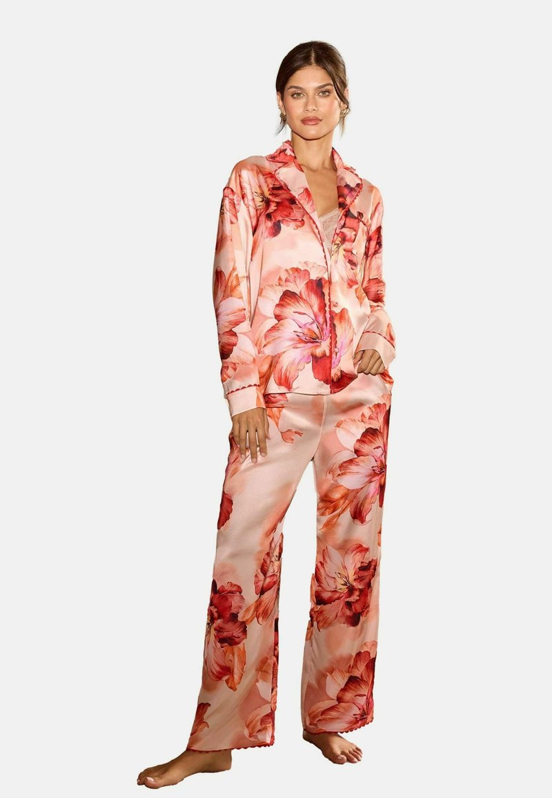 Set de pijama din satin floral, compus dintr-o bluză cu mâneci lungi și nasturi și pantaloni asortați, ambele de culoare roz deschis, cu flori mari roșii și portocalii.