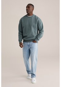 Klasičen zeleni pulover z rebrastimi manšetami in spodnjim robom, kombiniran s svetlo modrimi kavbojkami in belimi supergami. Preprost dizajn, sproščen kroj.