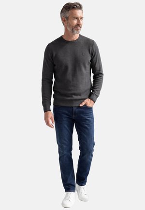 Mies, jolla on harmaat hiukset ja parta, pukeutuneena tummanharmaaseen neulepaitaan, sinisiin farkkuihin ja valkoisiin tennareihin, seisoo yksinkertaisen valkoisen taustan edessä.