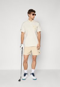 Beige polo-skjorte med brystlomme og broderte detaljer, kombinert med beige shorts. Tilbehør inkluderer solbriller og en golfklubbe.