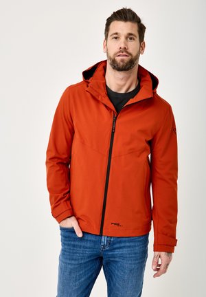 Mann mit Bart trägt eine orange Kapuzenjacke über einem schwarzen Shirt und blauen Jeans und steht vor einem weißen Hintergrund.