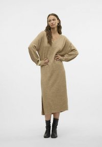 Robe tricotée beige avec décolleté en V, manches longues et fente sur le côté. Accentuée par des poignets côtelés. Associée à des bottes noires jusqu'au genou.
