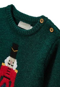 Pull en laine verte avec un motif de casse-noisette. Comprend un col rond, un tissu texturé et des boutons en bois sur l'épaule.