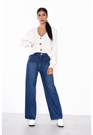 Crème kabelbrei cardigan met grote knopen, gecombineerd met hoge taille wijde blauwe jeans. Witte sneakers maken de outfit af.