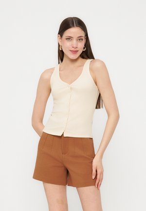 Γυναίκα που φοράει αμάνικη εκρού ριγέ μπλούζα με κουμπιά μπροστά και καφέ ψηλόμεσα, ραμμένα σορτς, στέκεται με το χέρι στη μέση πάνω σε λευκό φόντο.