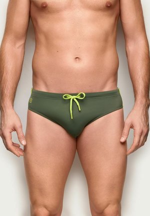 Buste masculin portant un slip de bain vert olive avec un cordon jaune néon, debout sur un fond clair uni.