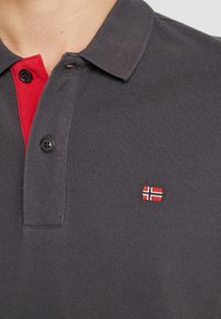 Polo gris foncé avec patte de boutonnage intérieure rouge, deux boutons noirs et un petit drapeau norvégien brodé sur la poitrine.