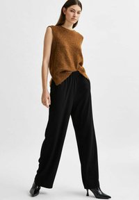 Haut en tricot sans manches marron avec un ourlet à franges associé à un pantalon large noir taille haute et des escarpins noirs brillants à bout pointu.