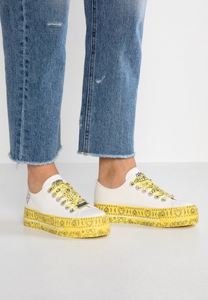 Baskets basses blanches avec semelles et lacets jaunes imprimés, portées avec un jean bleu court sur un fond uni.