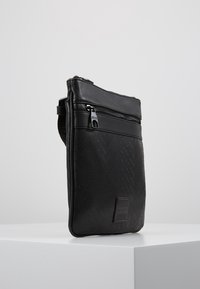Černá kabelka přes rameno z umělé kůže s herringbone texturou, zapínáním na zip a logem. Kompaktní obdélníkový tvar s nastavitelným popruhem.