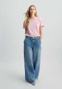 Tricou din bumbac roz deschis cu un logo mic pe piept, asortat cu blugi albaștri din denim, cu talie înaltă și croială lejeră. Textură netedă, croială casual.