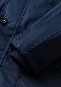 Chaqueta azul marino con textura suave, cierre de botones y puño de punto acanalado. Detalles de costura visibles en las mangas y el cuerpo.