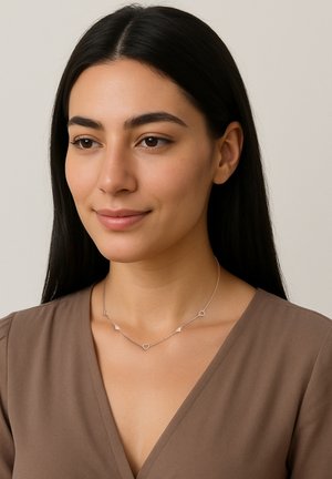 Una giovane donna con i capelli neri lisci indossa una delicata collana d'argento con piccoli ciondoli a forma di cuore e una maglietta marrone a scollo a V.