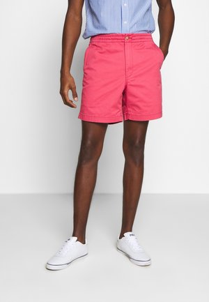 Man draagt roze korte broek, blauw-wit gestreept overhemd en witte sneakers, staand tegen een effen lichte achtergrond.