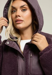 Fioletowa polarowa kurtka z kapturem, z zamykaną na zamek kieszenią, zapięciem na napy i miękką teksturą. Zawiera mieszankę gładkiego i puszystego materiału.
