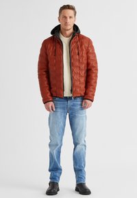 Braune, gesteppte Daunenjacke mit einer schwarzen Kapuze, kombiniert mit einem cremefarbenen Sweater und hellblauen Jeans. Schwarze Stiefeletten runden den Look ab.