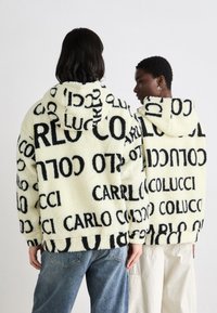 Fleece hoodie i lys gul med sort teksttryk. Har en hætte, lange ærmer og en afslappet pasform. Teksten lyder "CARLO COLUCCI" i fremtrædende skrift.