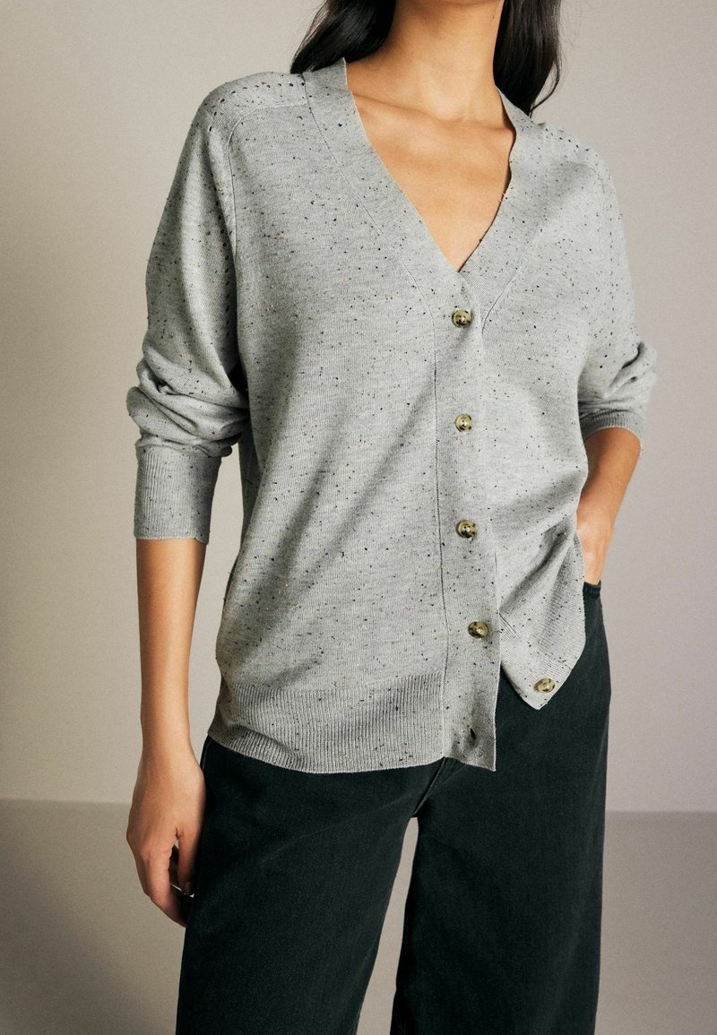 Cardigan grigio in morbido tessuto a maglia con scollatura a V, dotato di grandi bottoni dorati, polsini arrotolati e texture screziata. Abbinato a pantaloni scuri.