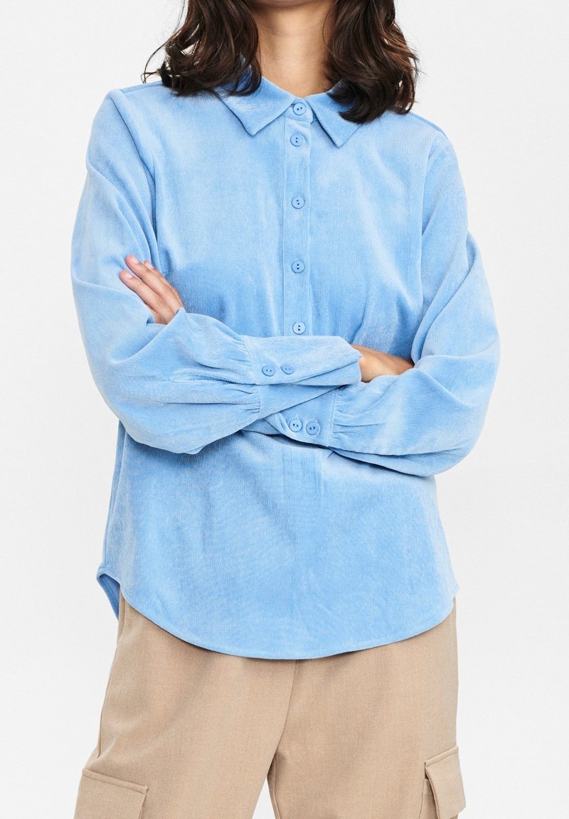 Camicia azzurra in velluto a coste con bottoni, colletto, maniche lunghe e tessitura sottile. Orlo arrotondato con polsini con bottoni.