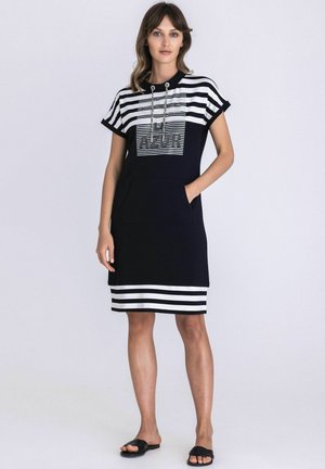 Femme debout portant une robe mi-longue rayée bleu marine et blanche avec manches courtes, poche avant, et sandales noires sans lacets.