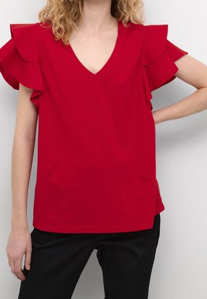 Vrouw met rode V-hals blouse met wijde mouwen en zwarte broek, staand met één hand op de heup tegen een effen witte achtergrond.