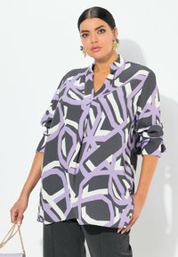 Schwarz und lavendelfarbene gemusterte Bluse mit langen Ärmeln, V-Ausschnitt und umgeschlagenen Manschetten, die abstrakte Formen in Weiß und Lila auf einem strukturierten Stoff zeigt.