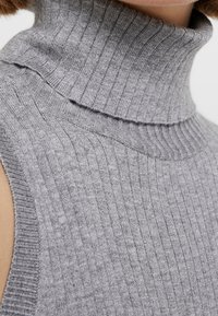 Grå ribbad polotröja med hög hals och en solid stickad textur. Modellen har en öppen axeldesign utan ärmar.