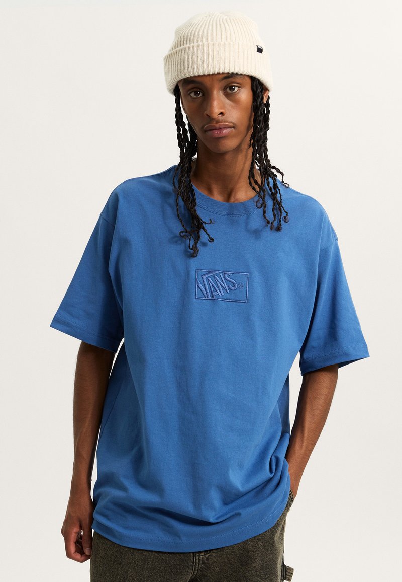 Camiseta de algodón azul con un logotipo de Vans bordado en el pecho. Combinada con un gorro blanco con canalé, el conjunto presenta un corte holgado y un estilo casual.