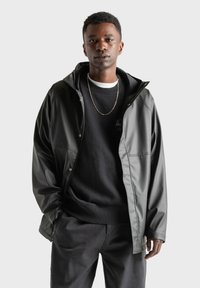 Zwarte waterdichte jas met een capuchon, gemaakt van gladde stof, met drukknoopsluiting en een relaxte pasvorm, gedragen over een zwart sweatshirt.