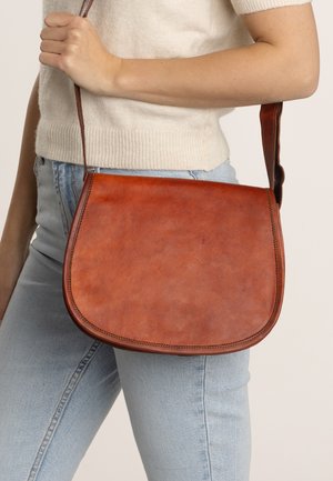 Hnědá kožená crossbody taška s kulatým klopou, hladkým povrchem a kontrastním prošíváním; držena v ruce, styling s světle modrými džíny.
