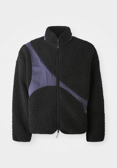 Svart fleecejakke med skrå lilla nylon aksent, høy krage, glidelås lukking og tekstil som gir varme.