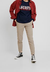 Röd jacka, marinblå sweatshirt med "Lacoste"-logotyp, beige rutiga byxor, svarta högst uppe sneakers med vita detaljer. Avslappnad ensemble.