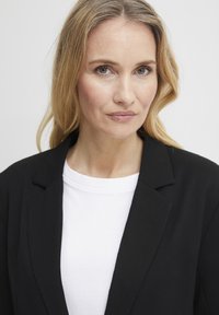 Črna blazer jakna čez belo majico. Blazer ima strukturirano zasnovo z zareznimi reverji. Tkanina izgleda gladka z nežen leskom.
