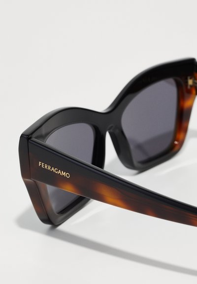 Occhiali da sole oversized neri e tartarugati con lenti grigie. Presentano la scritta oro "FERRAGAMO" sulla tempia. Finitura opaca e design angolare.